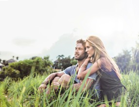 Ein junges Paar sitzt aneinander geschmiegt auf einer Wiese und blickt in die Ferne
